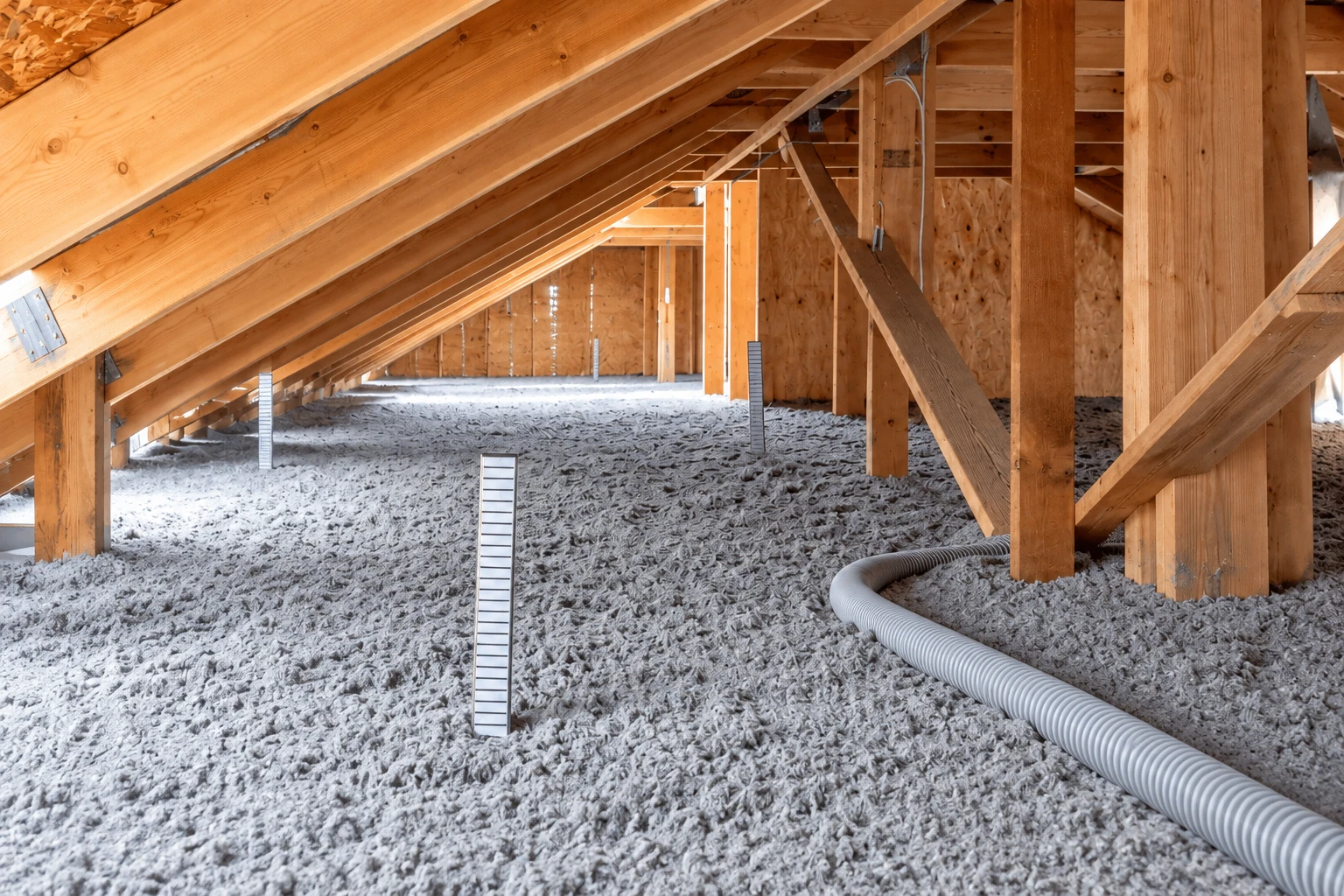 Gray blown-in insulation covers an attic floor beneath exposed wooden rafters, with a flexible hose in the foreground.