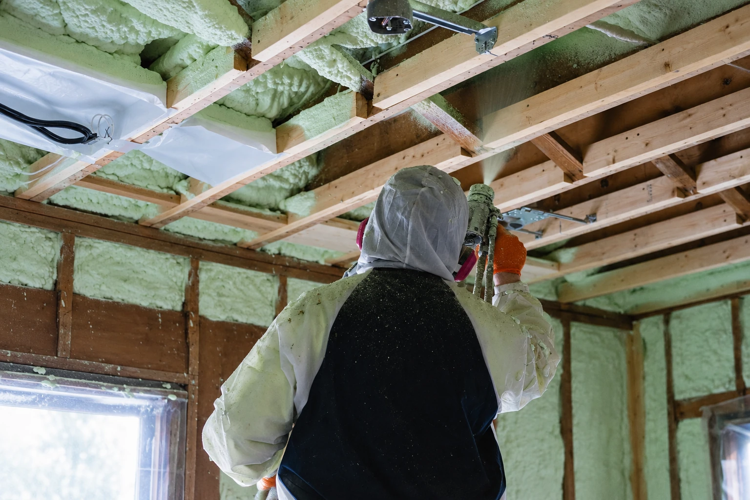 Worker in protective gear sprays pale green foam insulation into exposed ceiling cavities.
