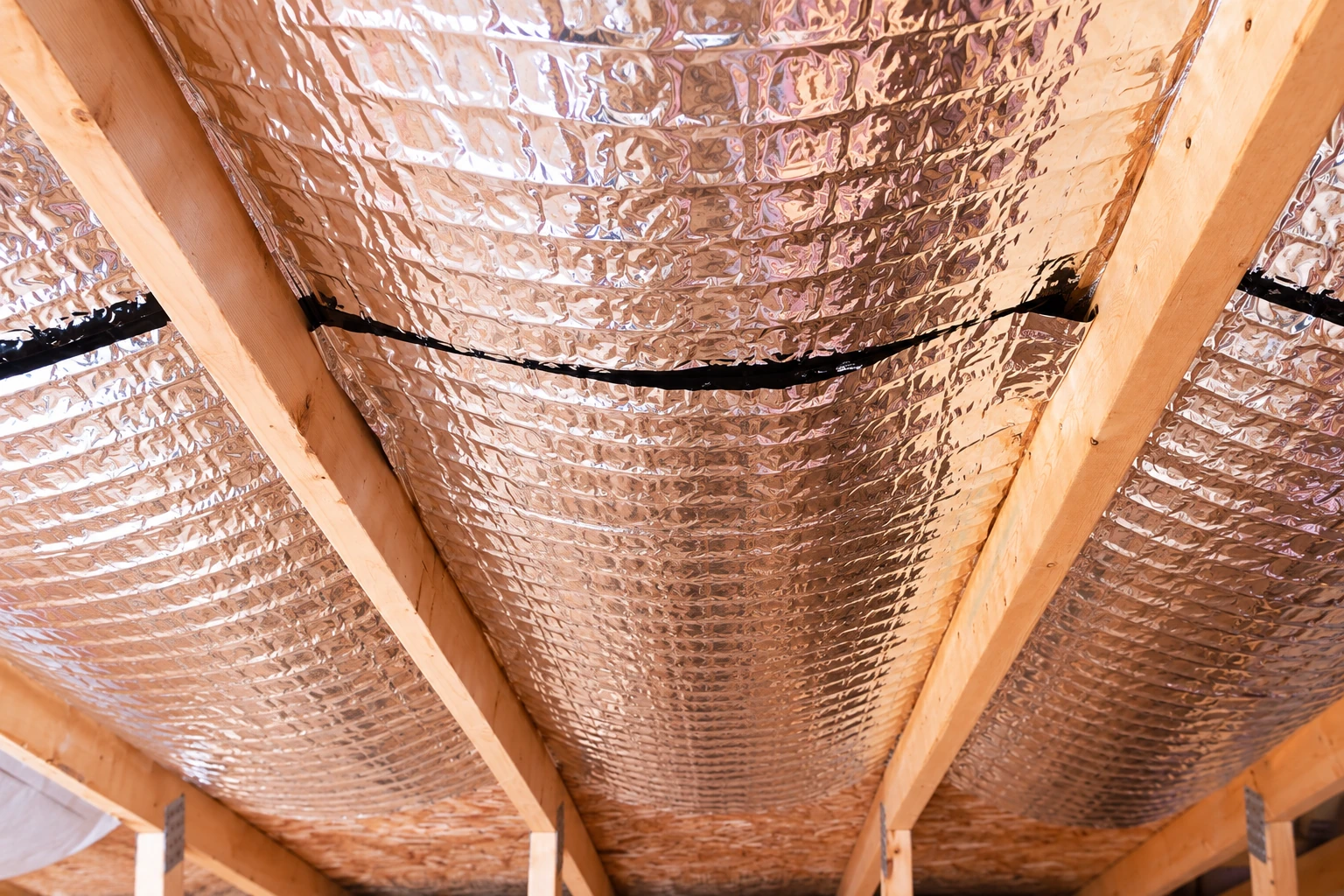 Reflective foil vapor barrier is fastened between wooden roof rafters, with a dark sealed seam running across it.